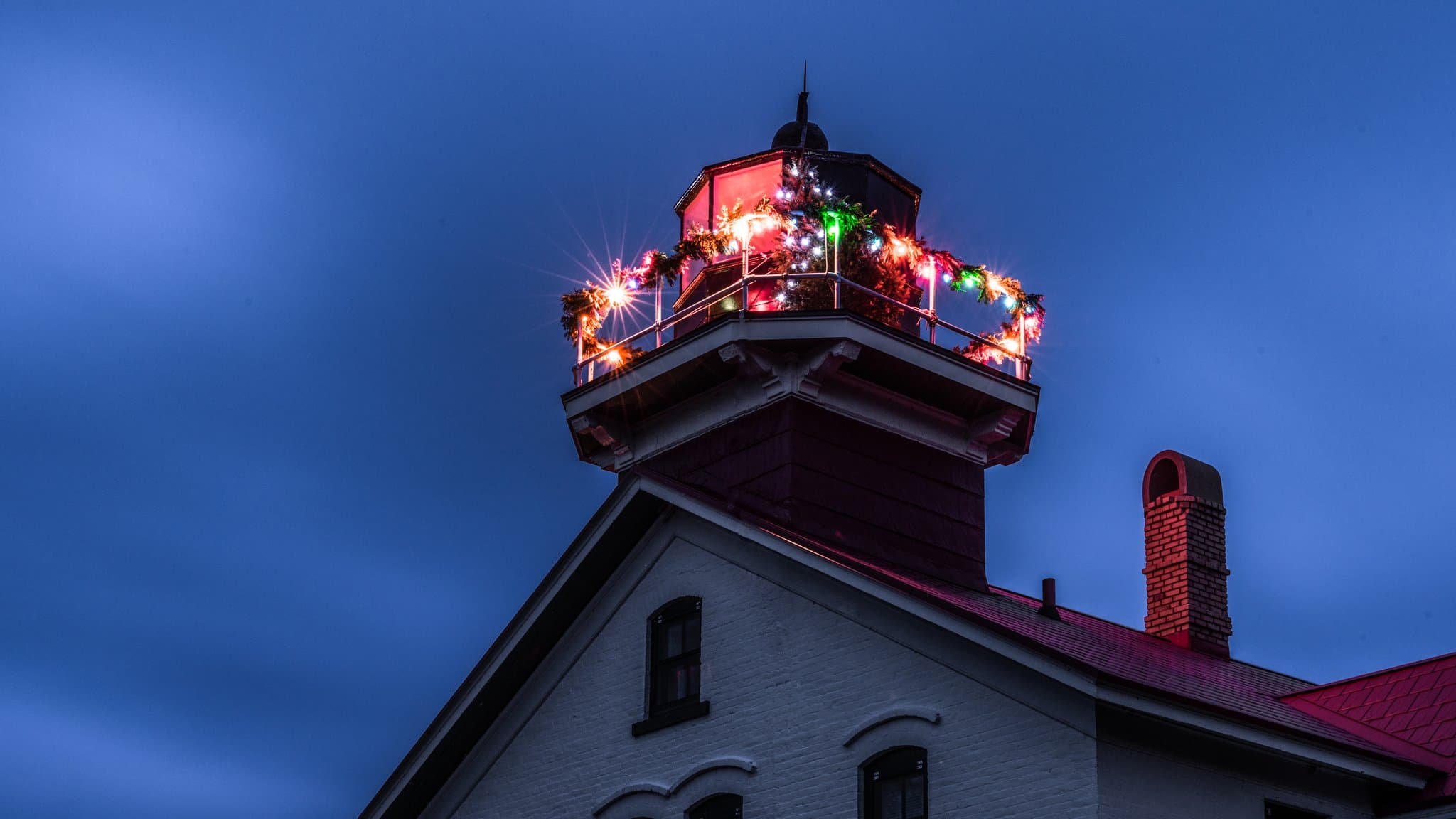 The Grand Traverse Lighthouse tower in Northport trimmed with colorful holiday lights and garland against a deep blue evening sky